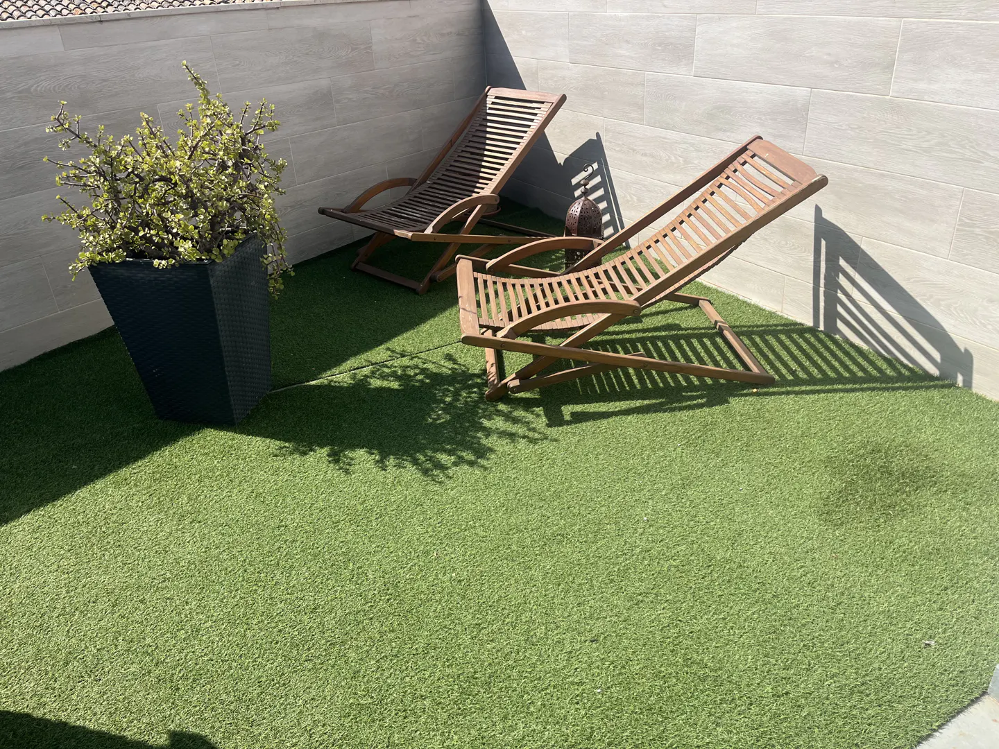 Outdoor patio with two wooden lounge chairs on green artificial turf, a black planter with a green plant, and a gray tiled wall.
