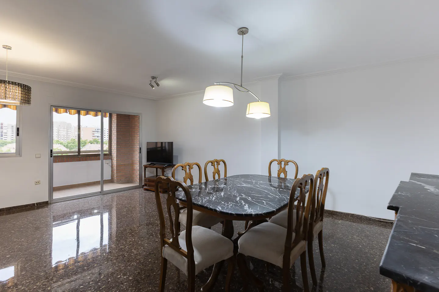 Bright dining room with marble table, six chairs, and a modern light fixture. Balcony with city view in the background.