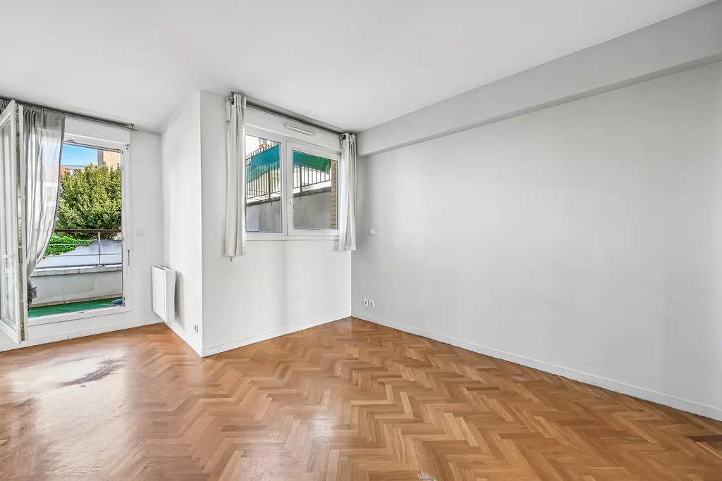 Bright, empty room with herringbone wood floors, white walls, and two windows. One window opens to a balcony with greenery.