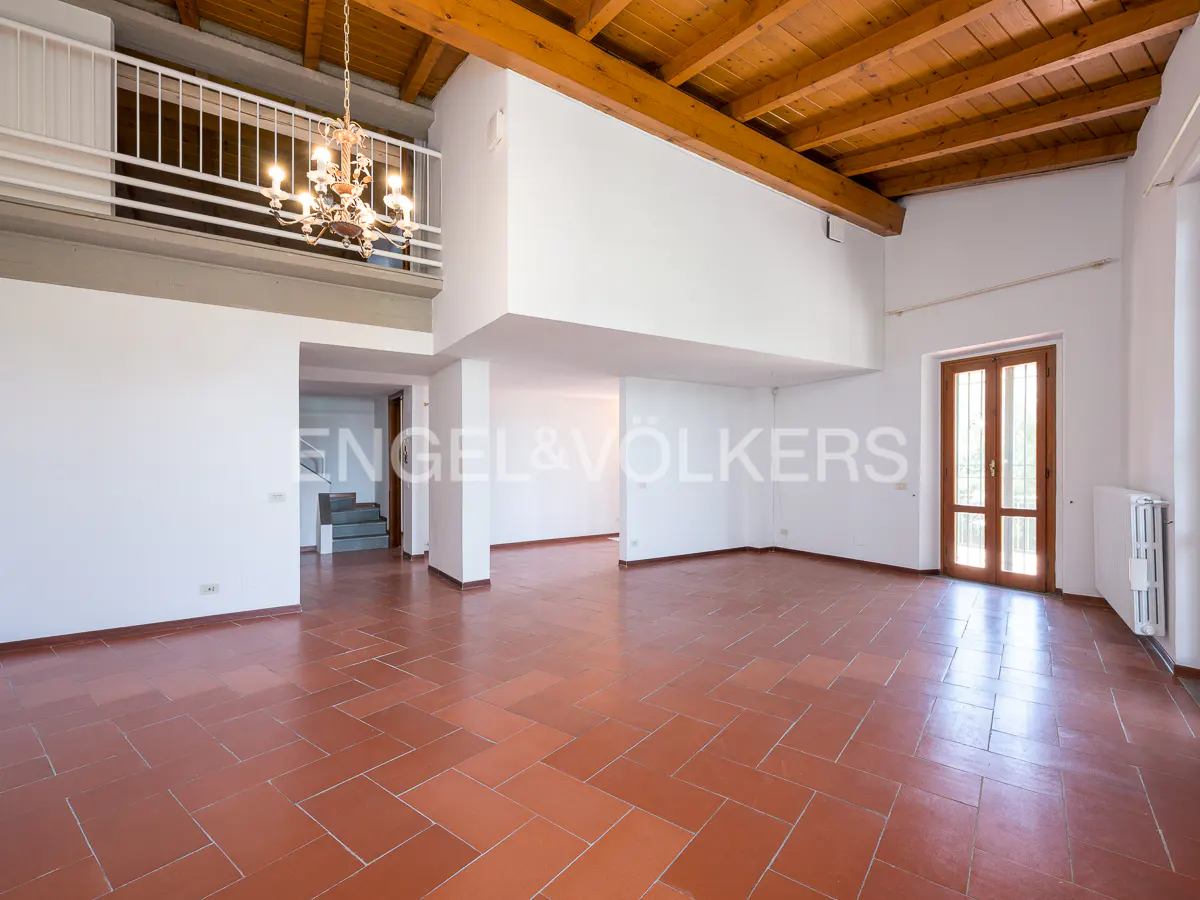 Spacious room with terracotta tile floor, white walls, and wood-beamed ceiling. A chandelier hangs from a balcony above.