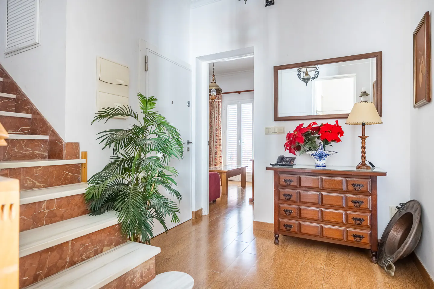 Bright entryway with marble stairs, a potted palm, a wooden chest of drawers with a lamp, and a view into a living room.