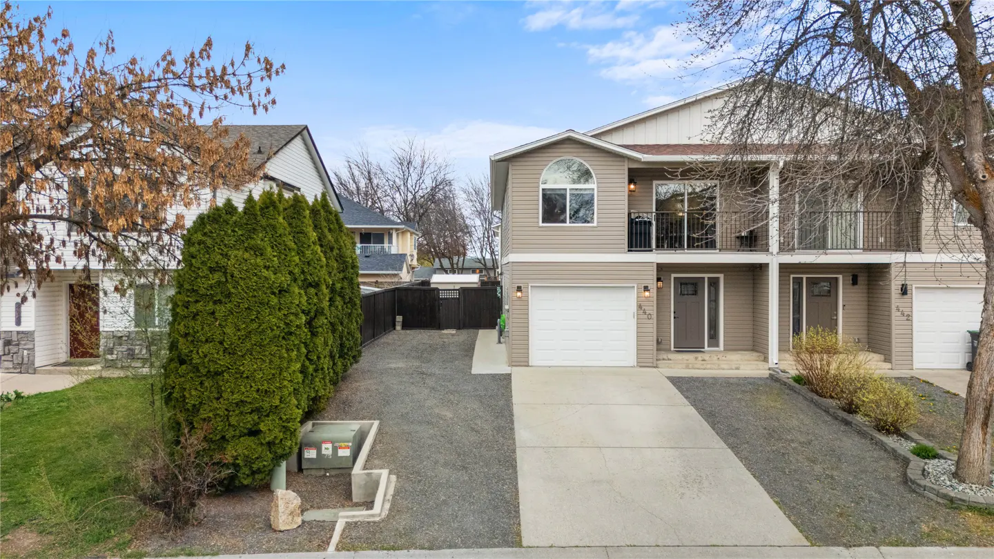 Tan two-story townhouse with white trim, garage, and small balcony. A row of green trees lines the driveway. Blue sky.