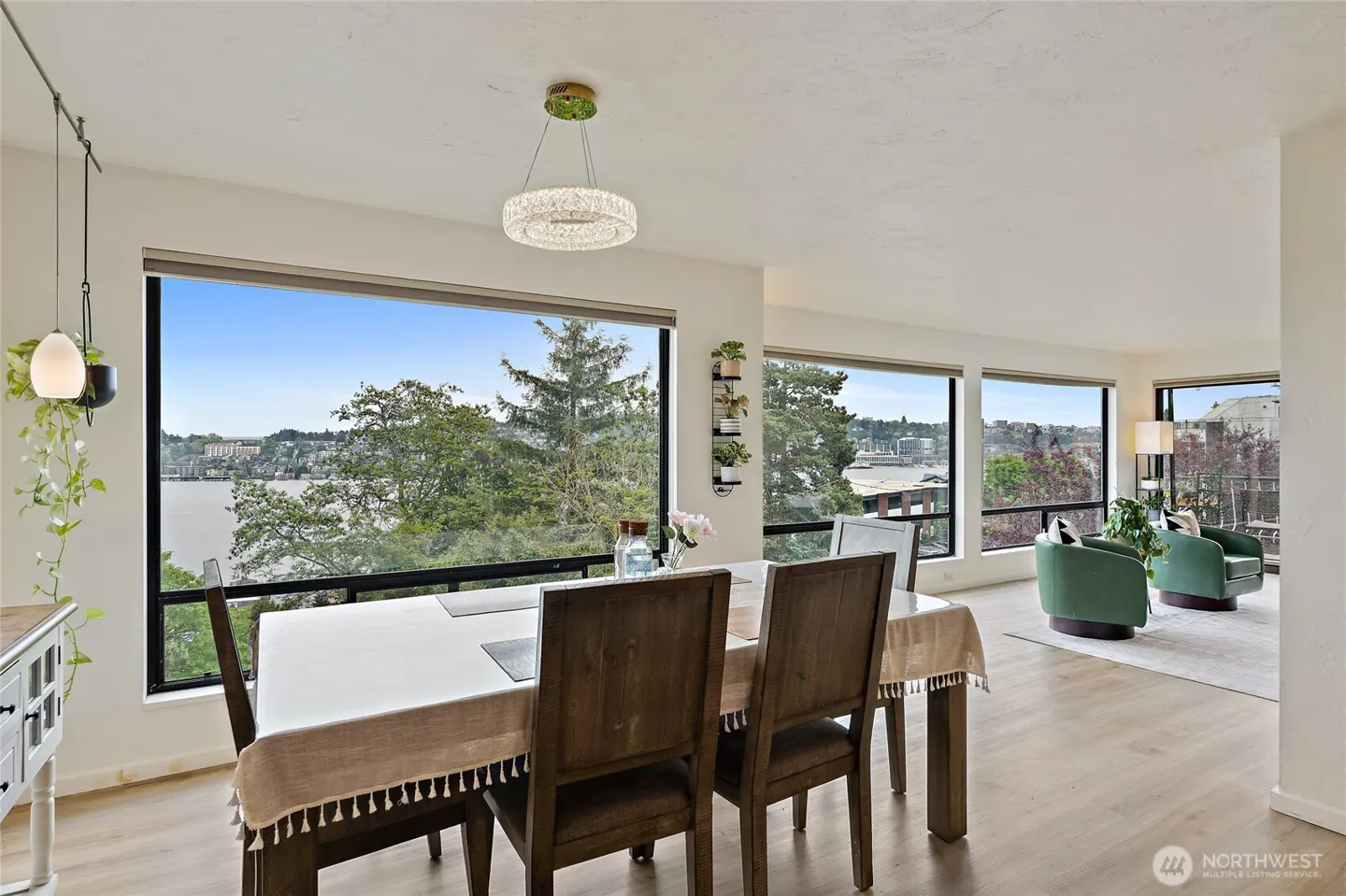 Bright dining room with wood table, chairs, and large windows overlooking a lake and trees. Green armchairs are visible in the background.