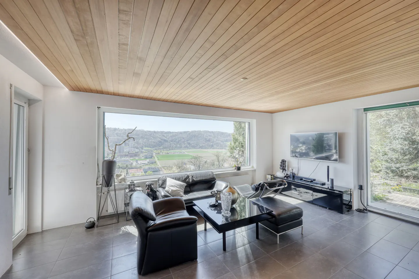 Bright living room with wood ceiling, gray tile floor, black leather furniture, and a large window with a view.