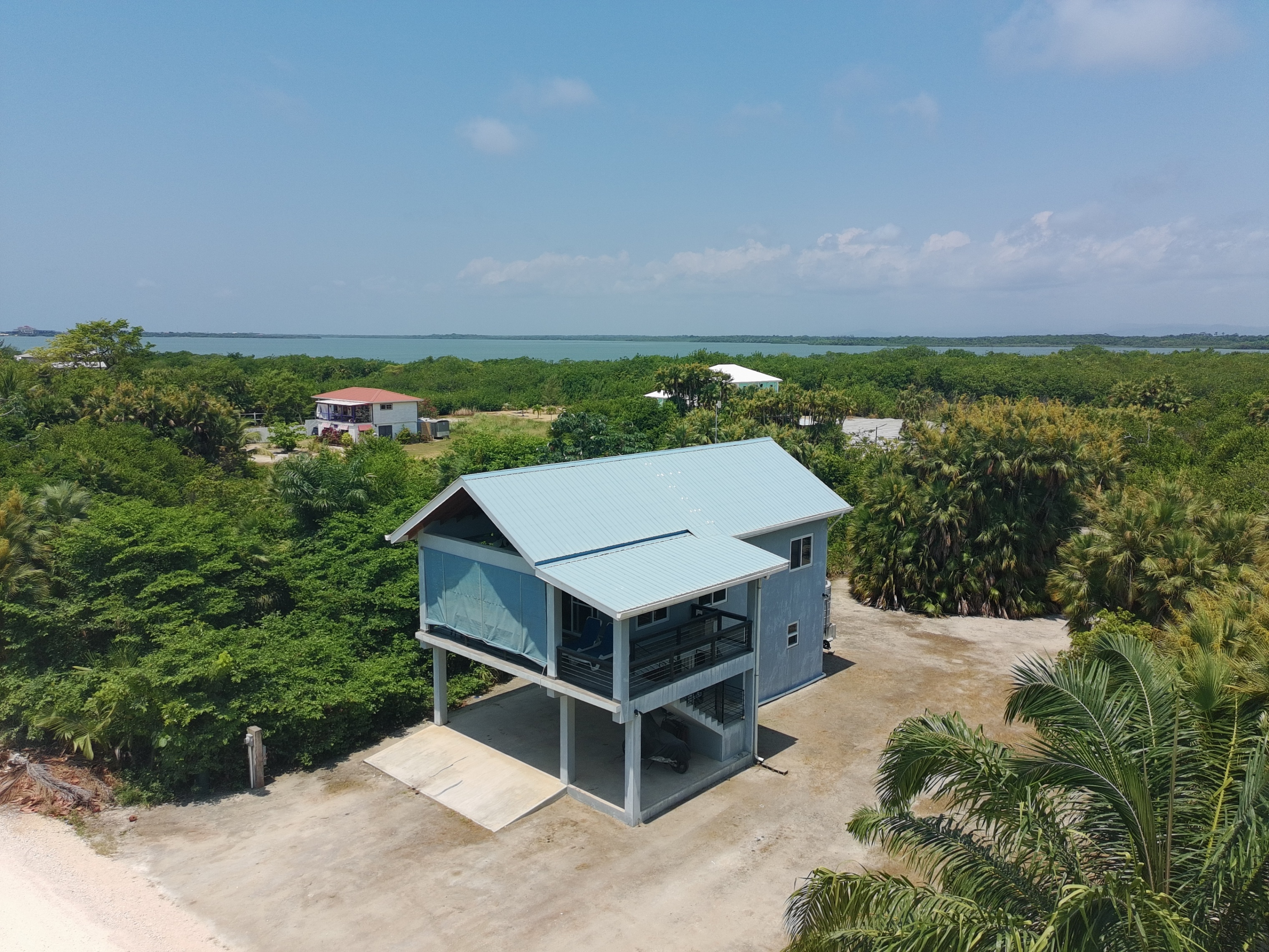 Two-story stilt house with a light blue roof and siding, surrounded by lush green trees and a glimpse of the ocean in the background.