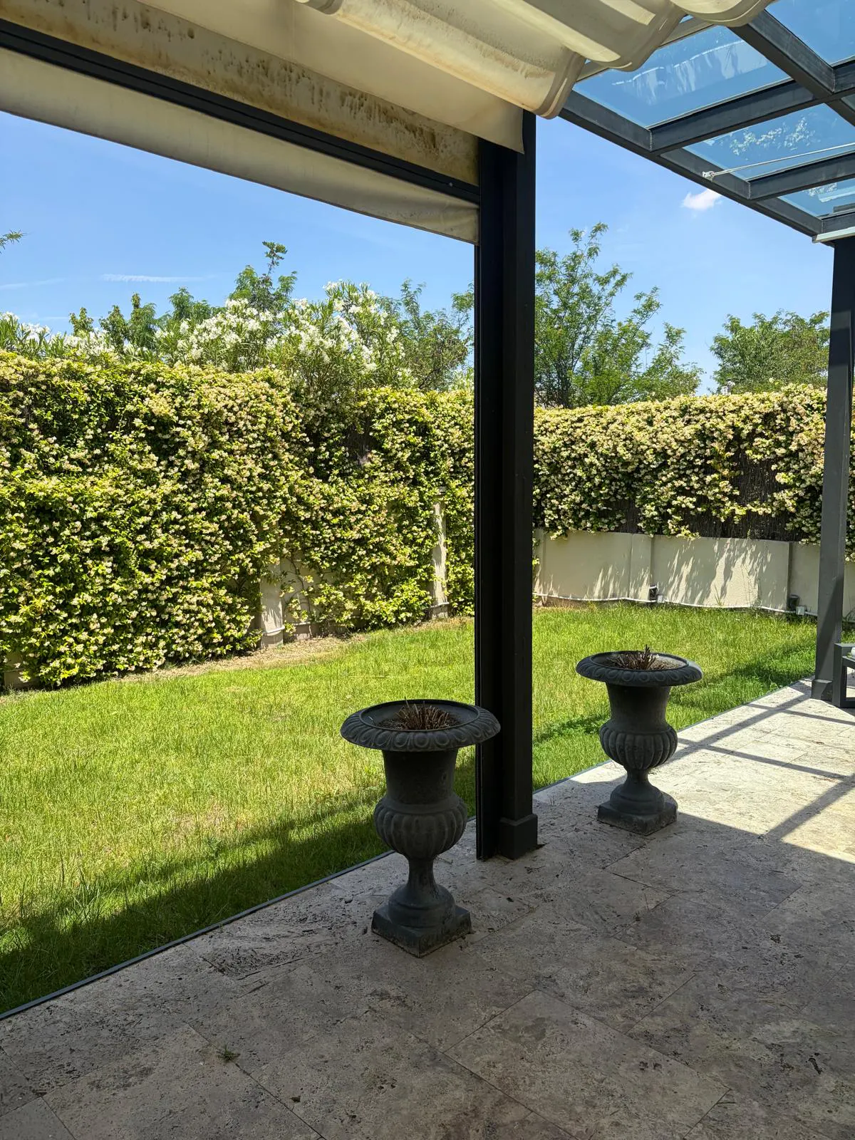 Outdoor patio with two gray urns, green lawn, and tall hedge under a blue sky.