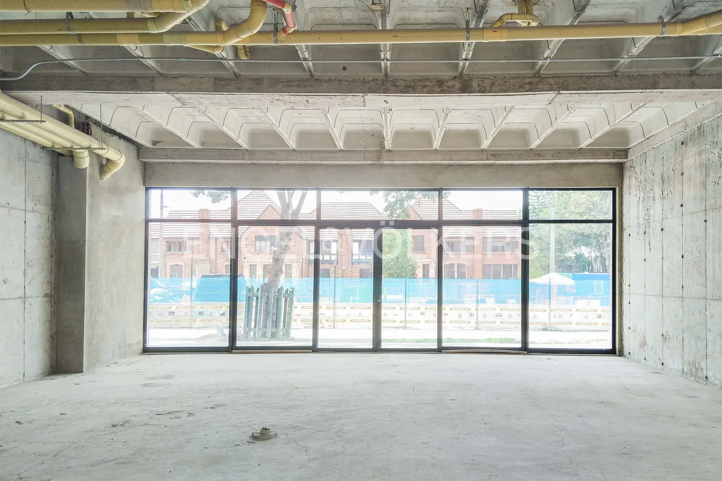 Unfinished commercial space with concrete walls, exposed ceiling pipes, and a large window overlooking a building.