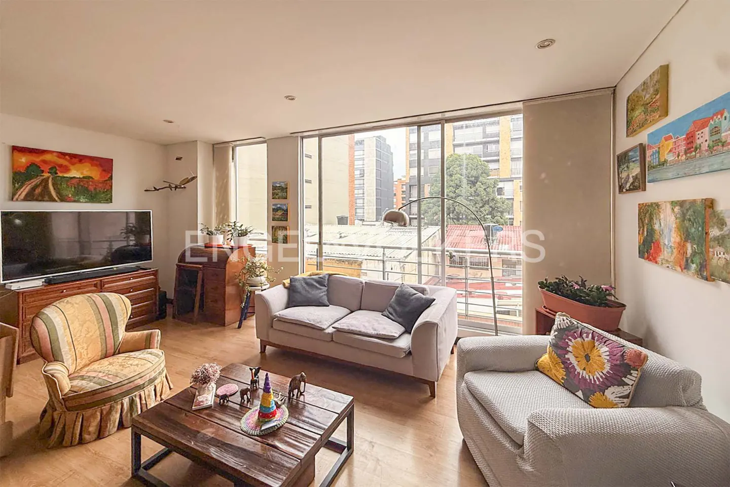 Bright living room with hardwood floors, a gray sofa, and a striped armchair. A large window overlooks city buildings. Artwork adorns the walls.