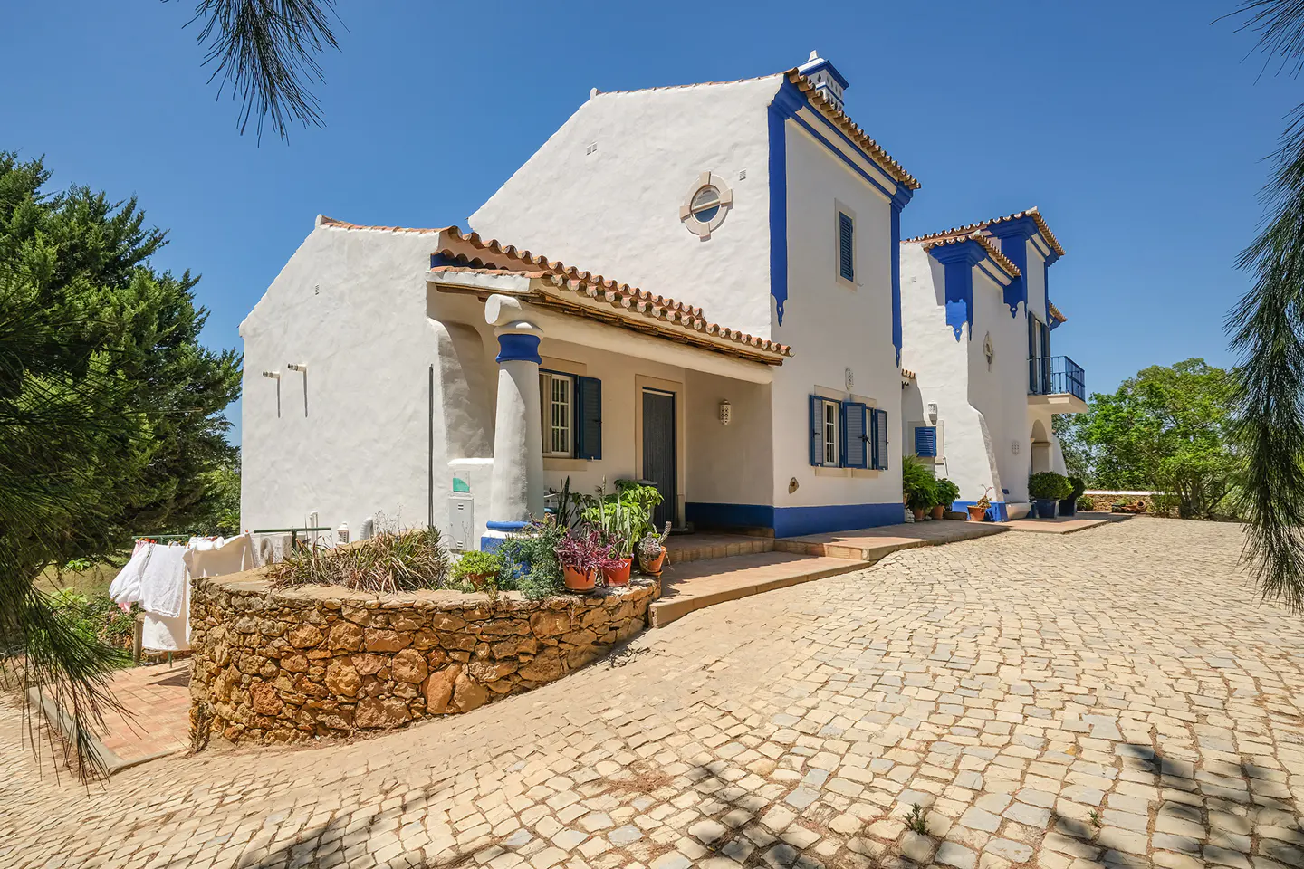 Two-story white house with blue trim and terracotta roof tiles on a cobblestone driveway under a clear blue sky.