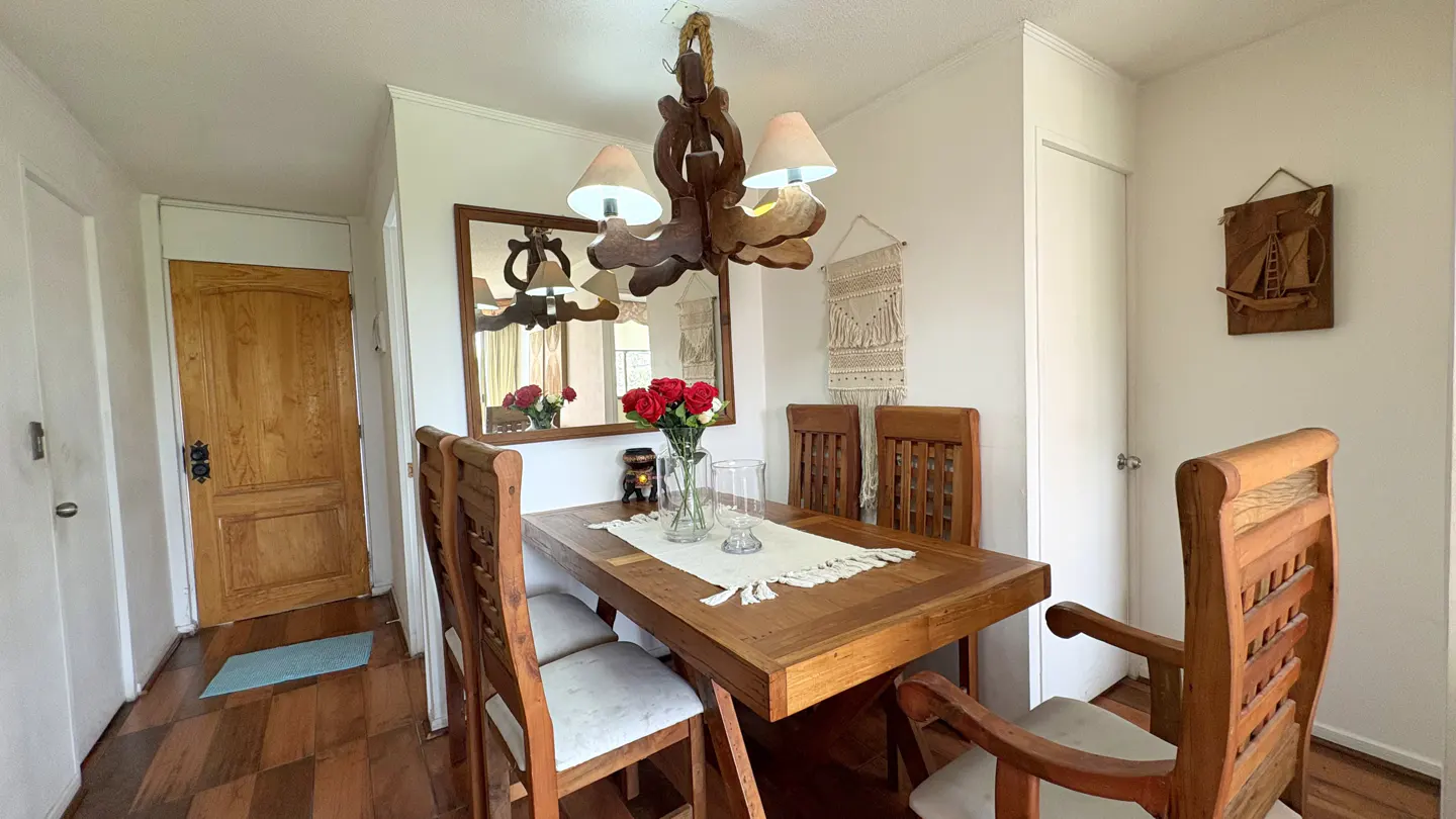 A dining room with a wooden table, chairs, and a unique wooden chandelier. Red roses in a vase add a touch of color. A mirror and wall art decorate the white walls.