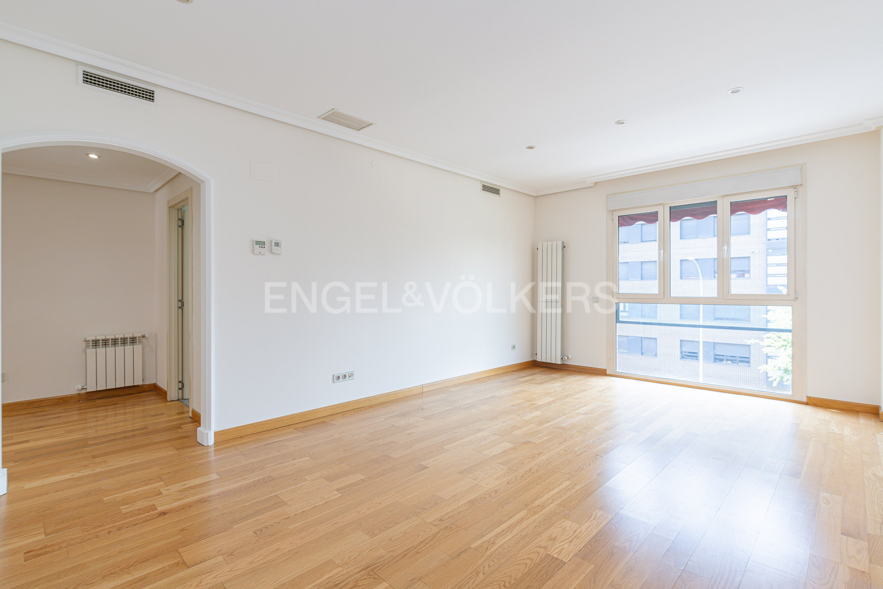 Bright, empty room with light wood floors, white walls, a large window, and an arched doorway leading to a hallway. [AI-generated text]