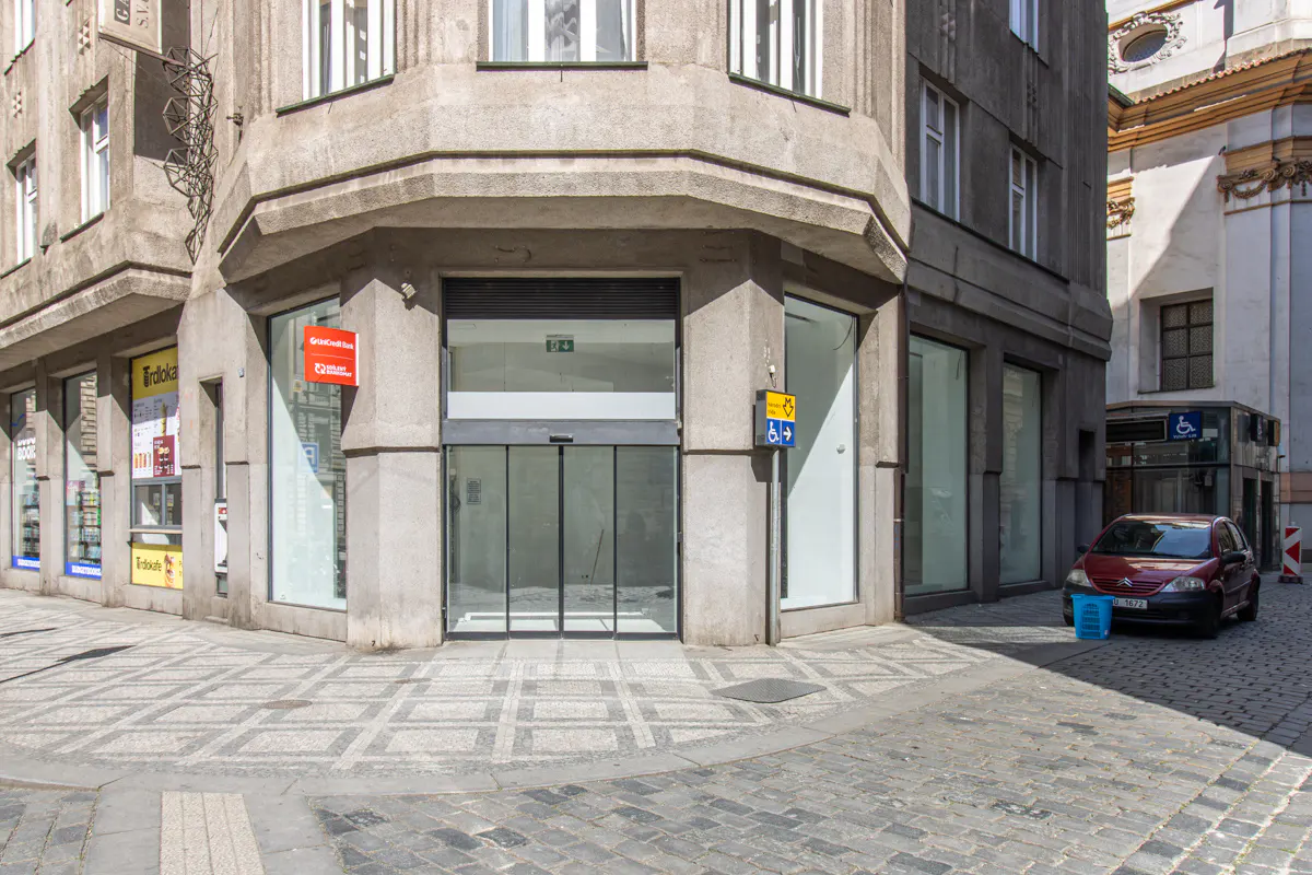 Corner building with glass storefront and automatic doors. A red car is parked on the cobblestone street.