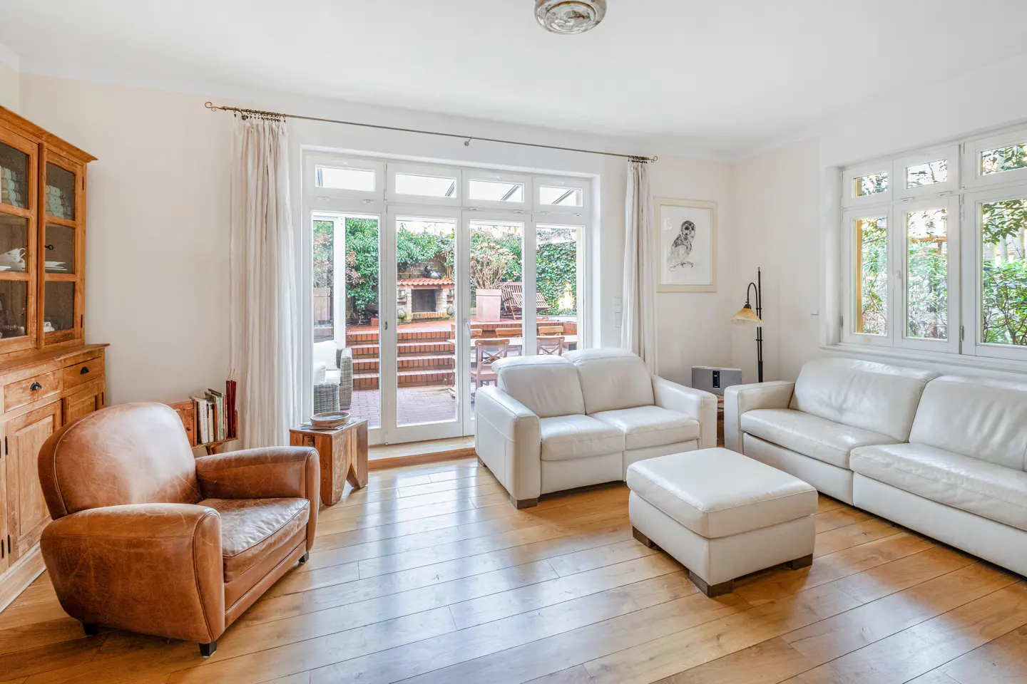 Bright living room with hardwood floors, white sofas, and a brown leather armchair. French doors lead to a patio with greenery.