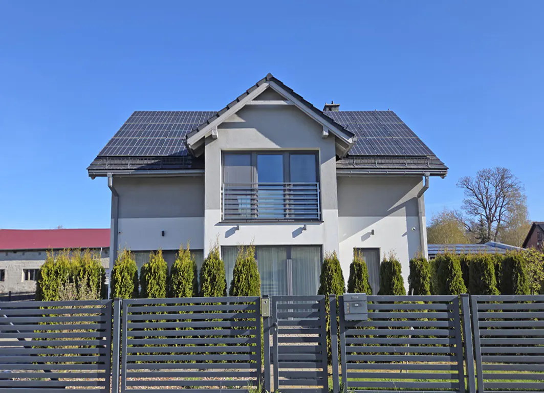 Two-story gray house with solar panels on the roof, a gray fence, and green bushes in front.