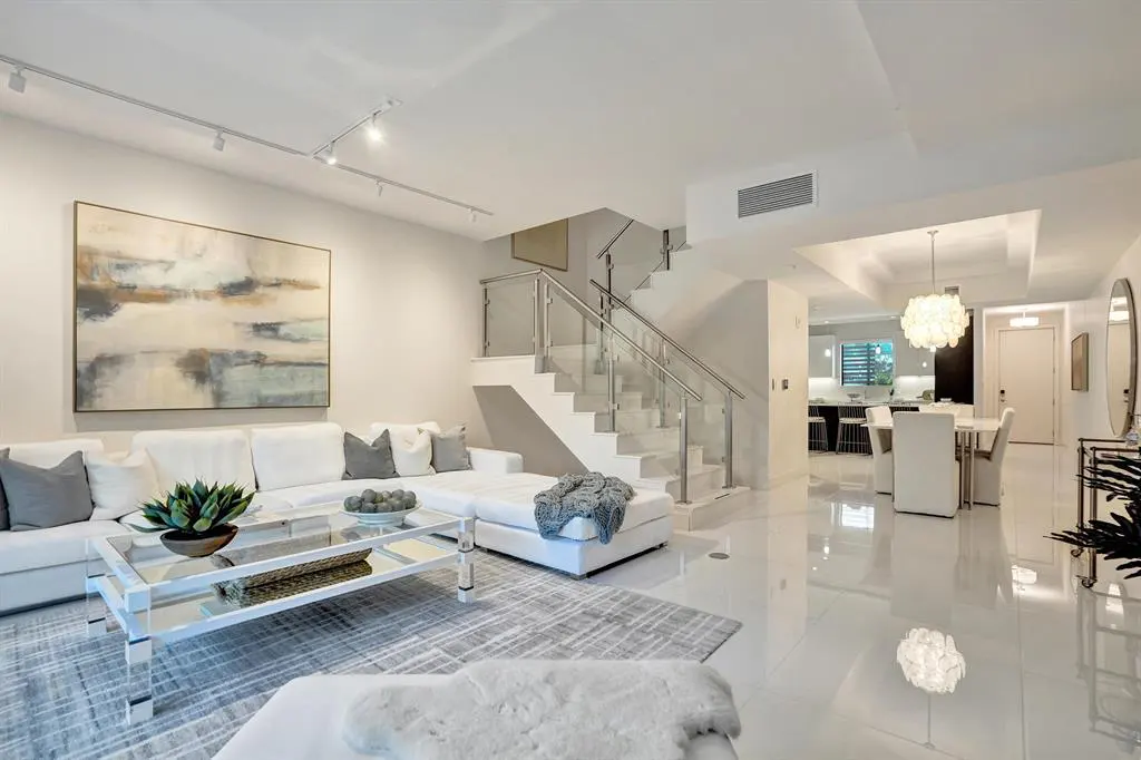 Bright, modern living room with white sectional sofa, glass coffee table, and abstract art. Staircase with glass railing leads upstairs.