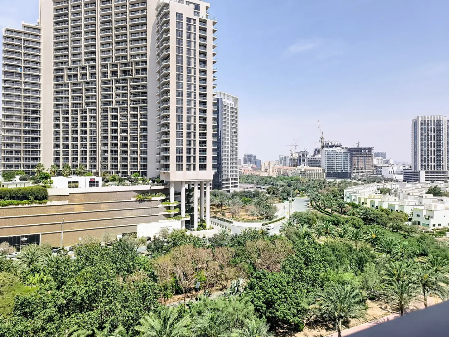 View of tall, modern apartment buildings with balconies, lush green trees, and other buildings in the background under a blue sky.