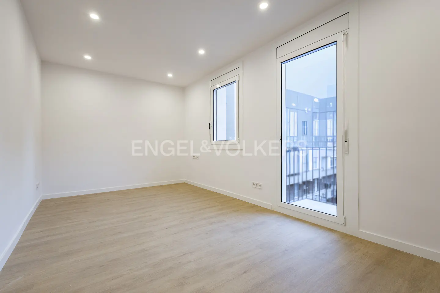 Bright, empty room with light wood floors, white walls, and recessed lighting. Large windows offer a view of a building. Engel & Volkers logo visible.