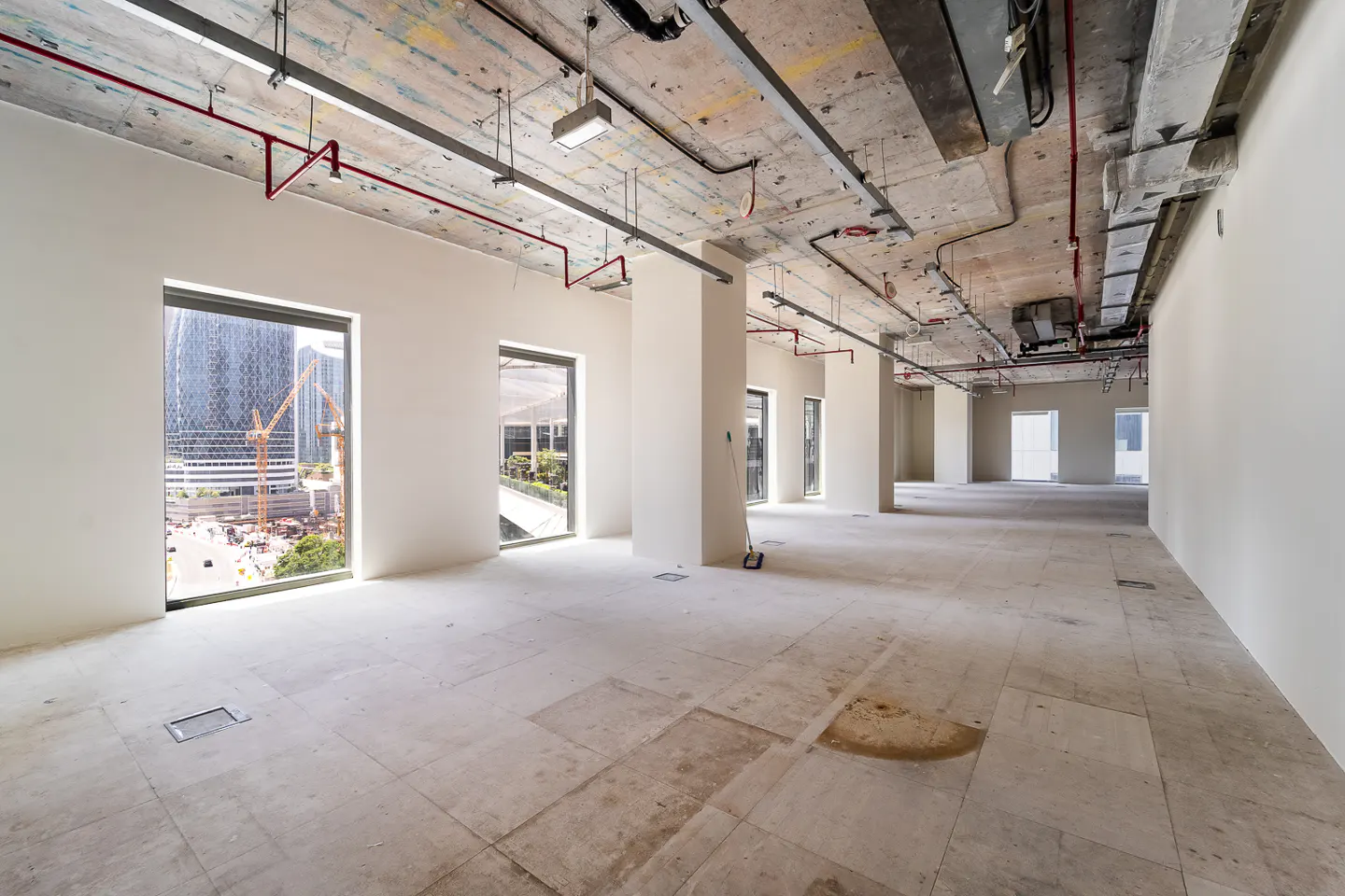 Unfinished office space with concrete floors and ceiling. Large windows offer city views. Red pipes and silver ducts run along the ceiling.