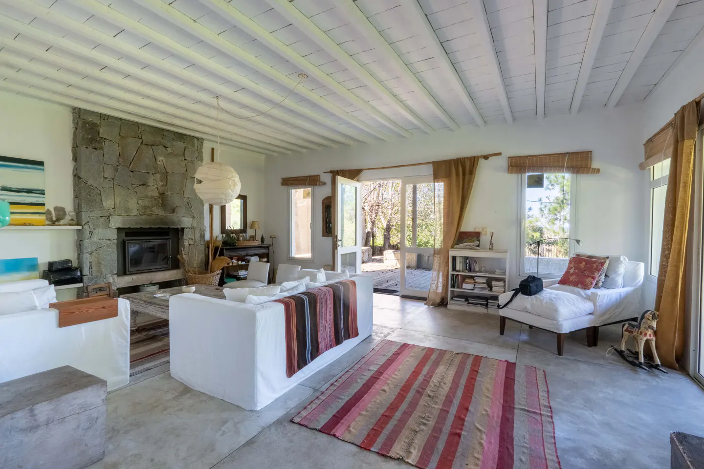 Bright living room with white sofas, stone fireplace, and striped rug. Open doors lead to an outdoor patio.