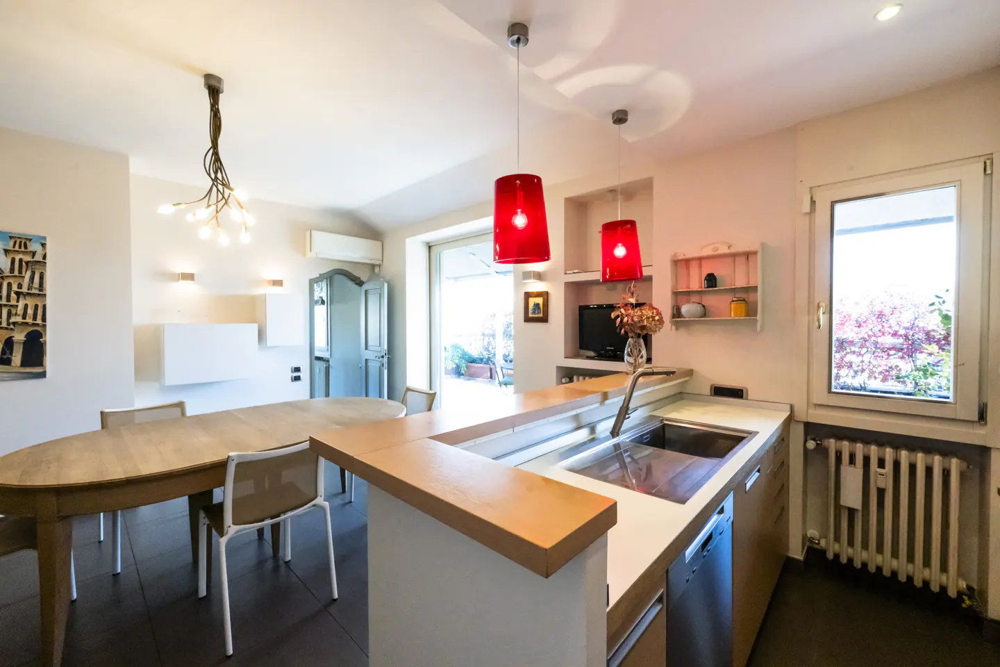 Bright, modern kitchen with a wooden table, chairs, and red pendant lights. A window shows a view of trees.
