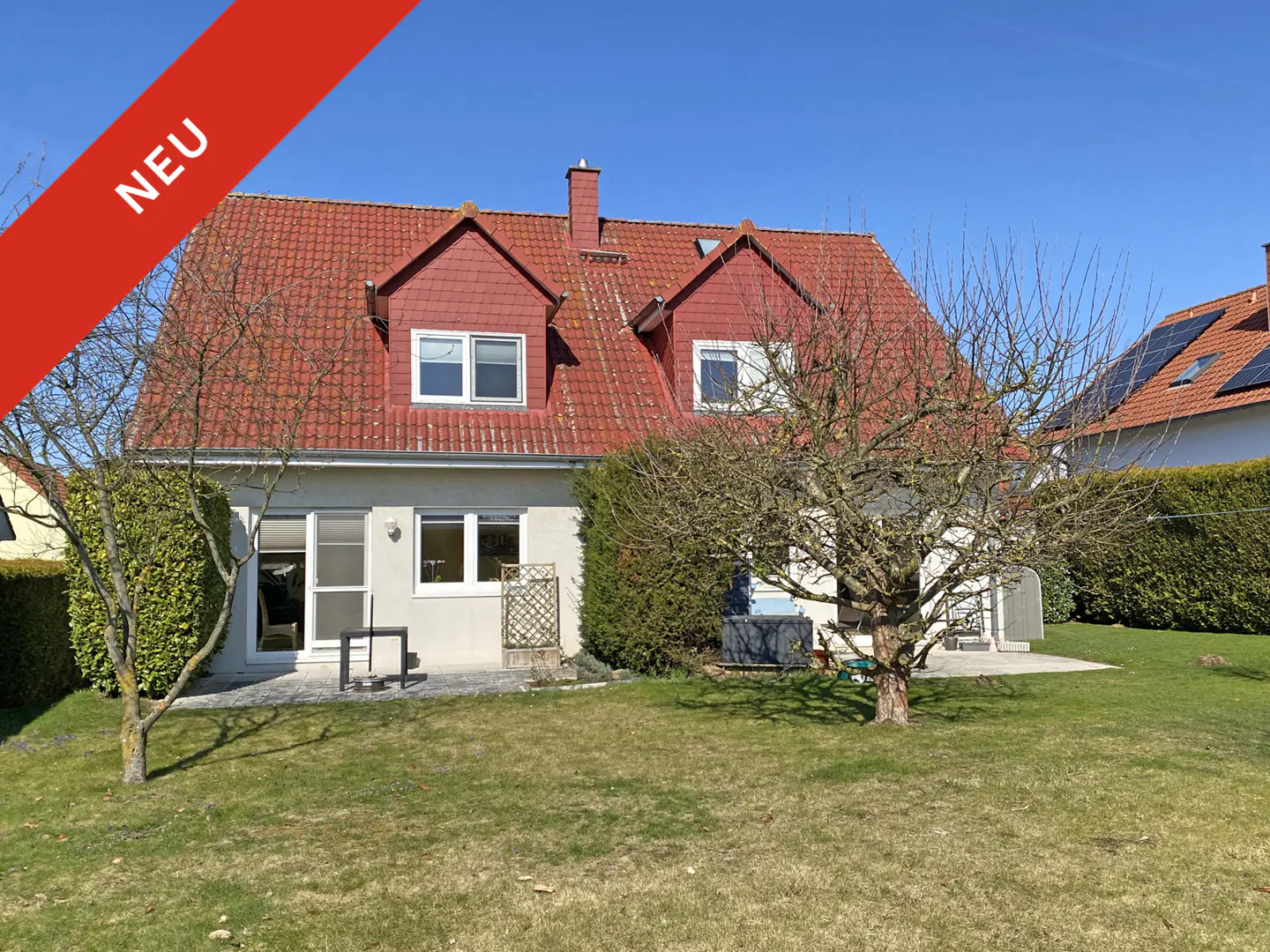 Two-story house with a red roof and a "Neu" banner, surrounded by a green lawn and trees under a clear blue sky.