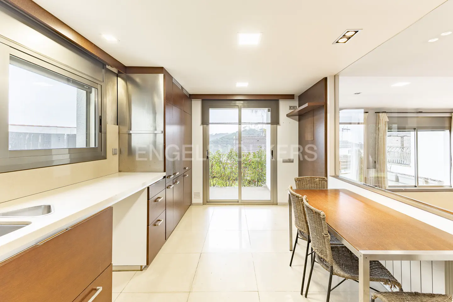A bright kitchen with wood cabinets, white countertops, and a dining table with wicker chairs. Sliding glass doors lead to a balcony with greenery.