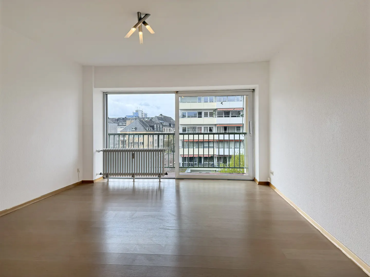 Empty room with white walls, wood floor, and a large window. A radiator sits below the window, with a city view visible outside.