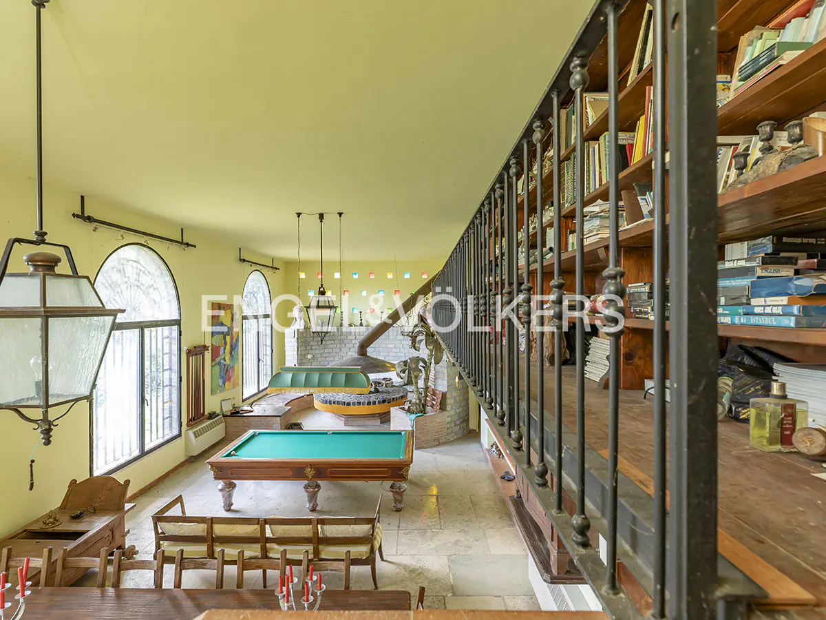 Interior view of a yellow room with a pool table, seating, and a bookshelf-lined staircase. Large windows provide natural light.