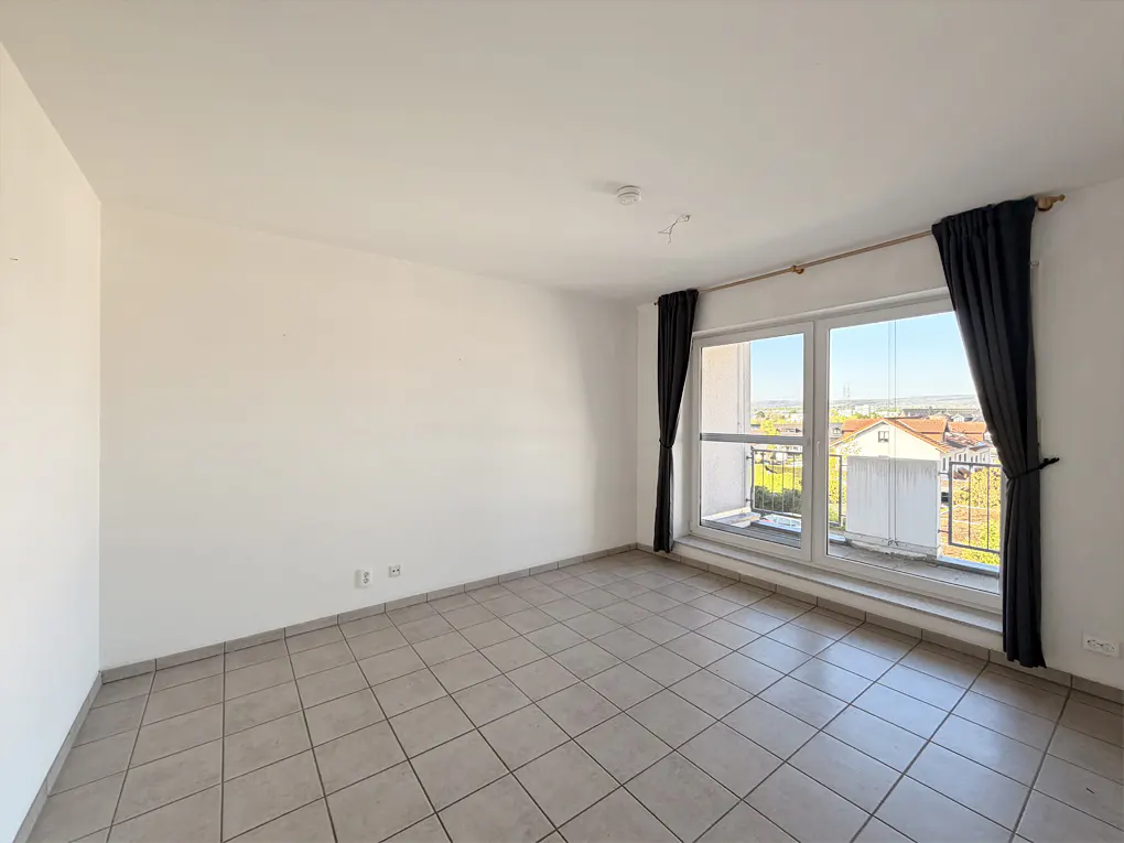 Bright, empty room with white walls, tile floor, and large window with black curtains. Balcony and cityscape visible outside.