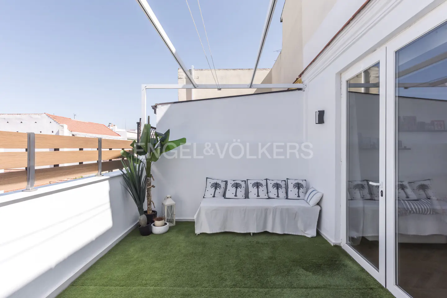 Bright rooftop terrace with artificial grass, a white bench with palm-print cushions, potted plants, and glass doors.