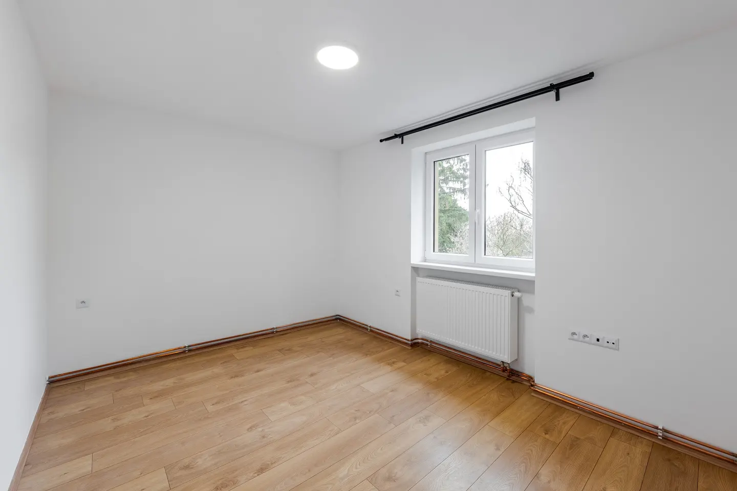 Bright, empty room with white walls, wood floor, and a window showing trees outside. A white radiator sits under the window.