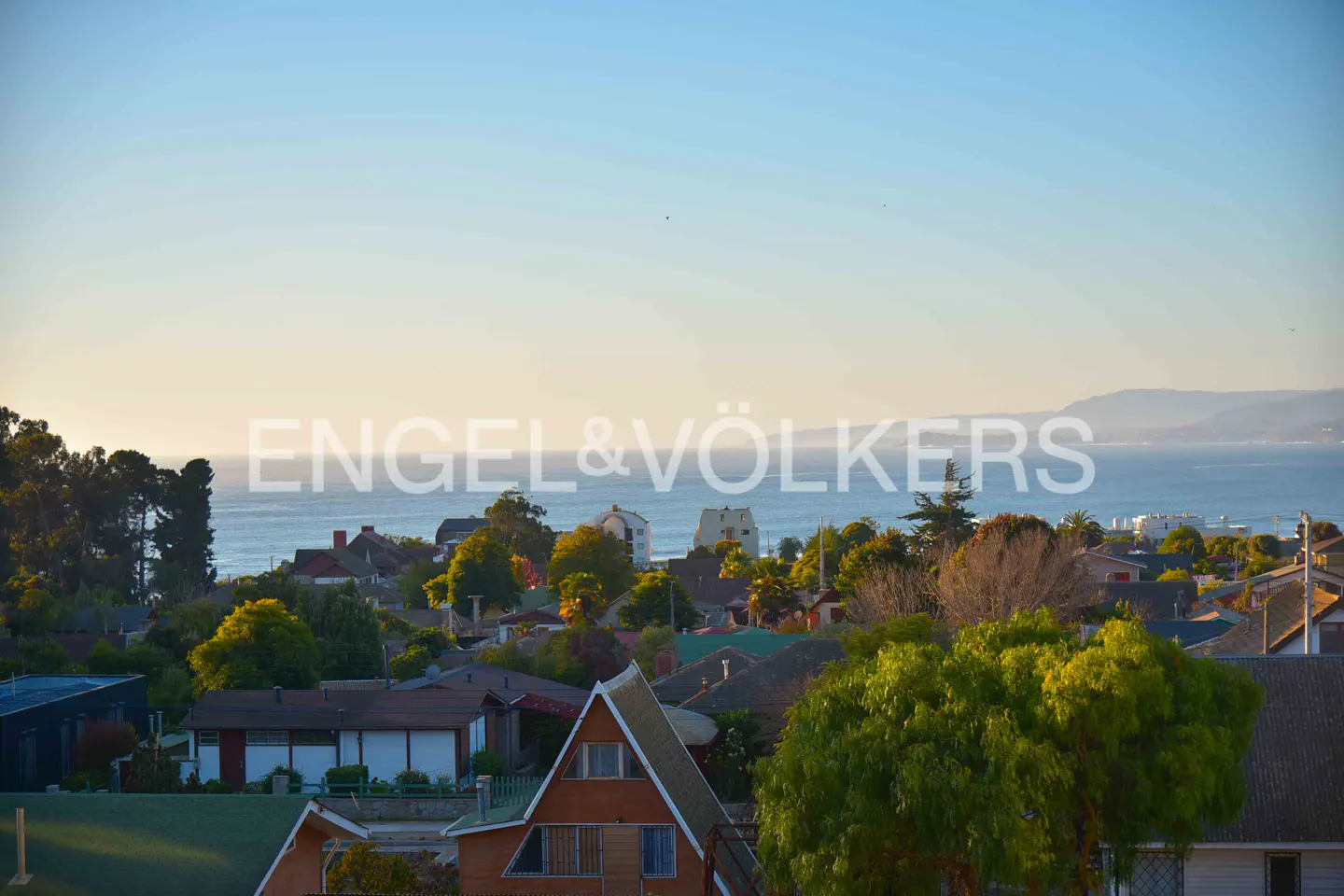 Scenic view of a coastal town with houses, trees, and the ocean under a clear blue sky.