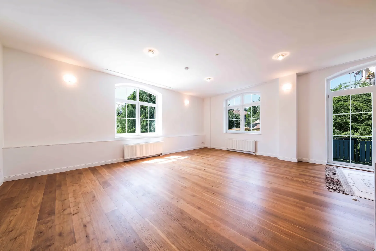 Bright, empty room with hardwood floors, white walls, and natural light from windows and a door to a balcony with green trees outside.