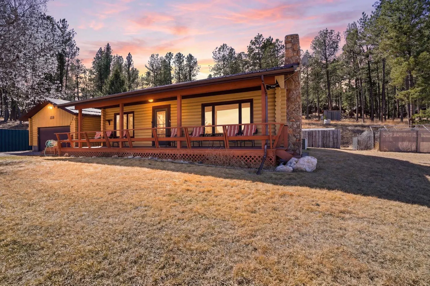 Exterior view of a log cabin home with a wooden porch, stone chimney, and a yard with dry grass. The sky is pink and orange.