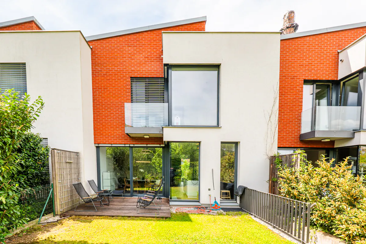 Modern townhouse with a brick and white facade, a wooden deck with chairs, and a green lawn.