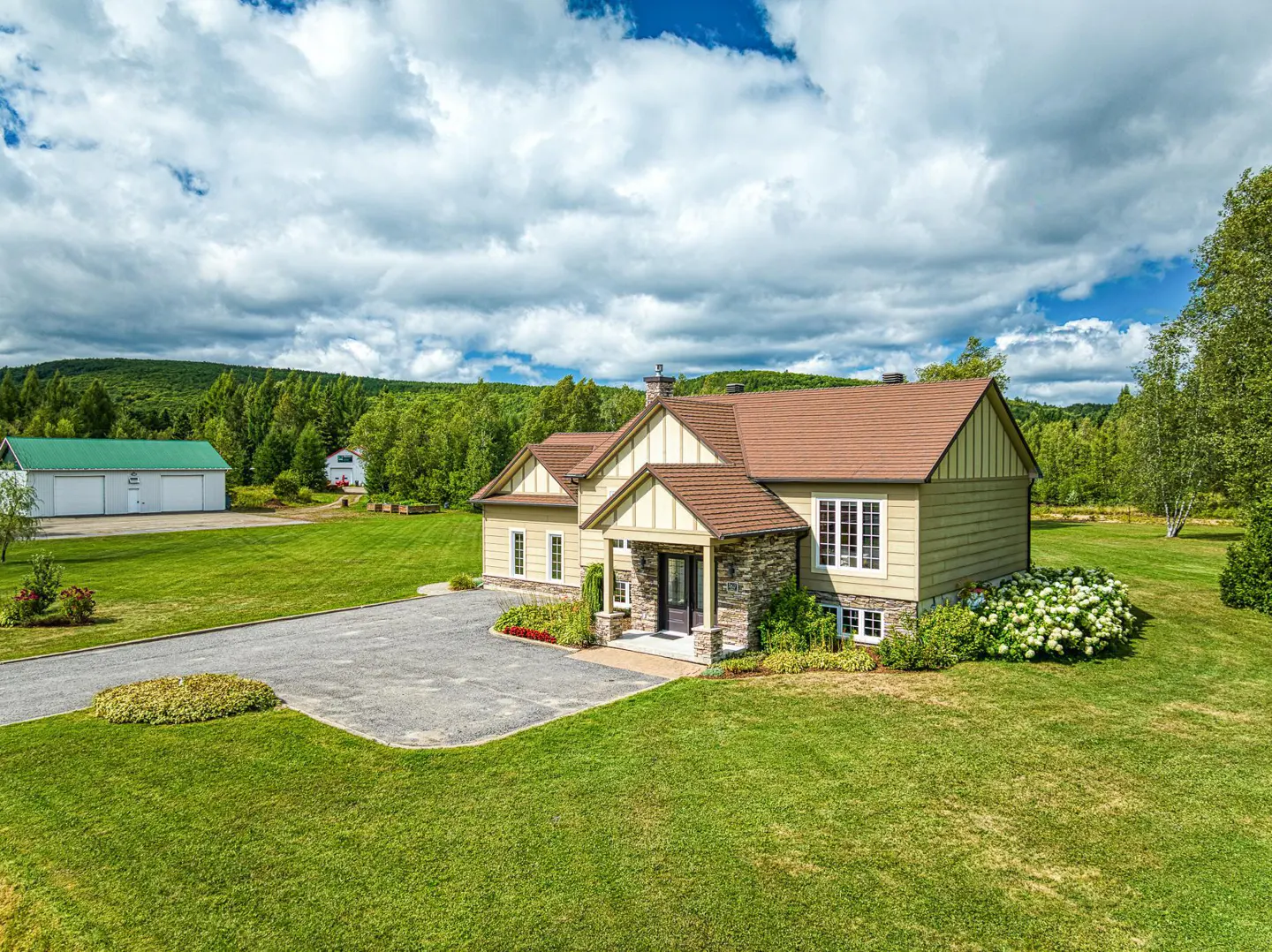 Tan house with brown roof, stone accents, and green lawn. A green barn is in the background.