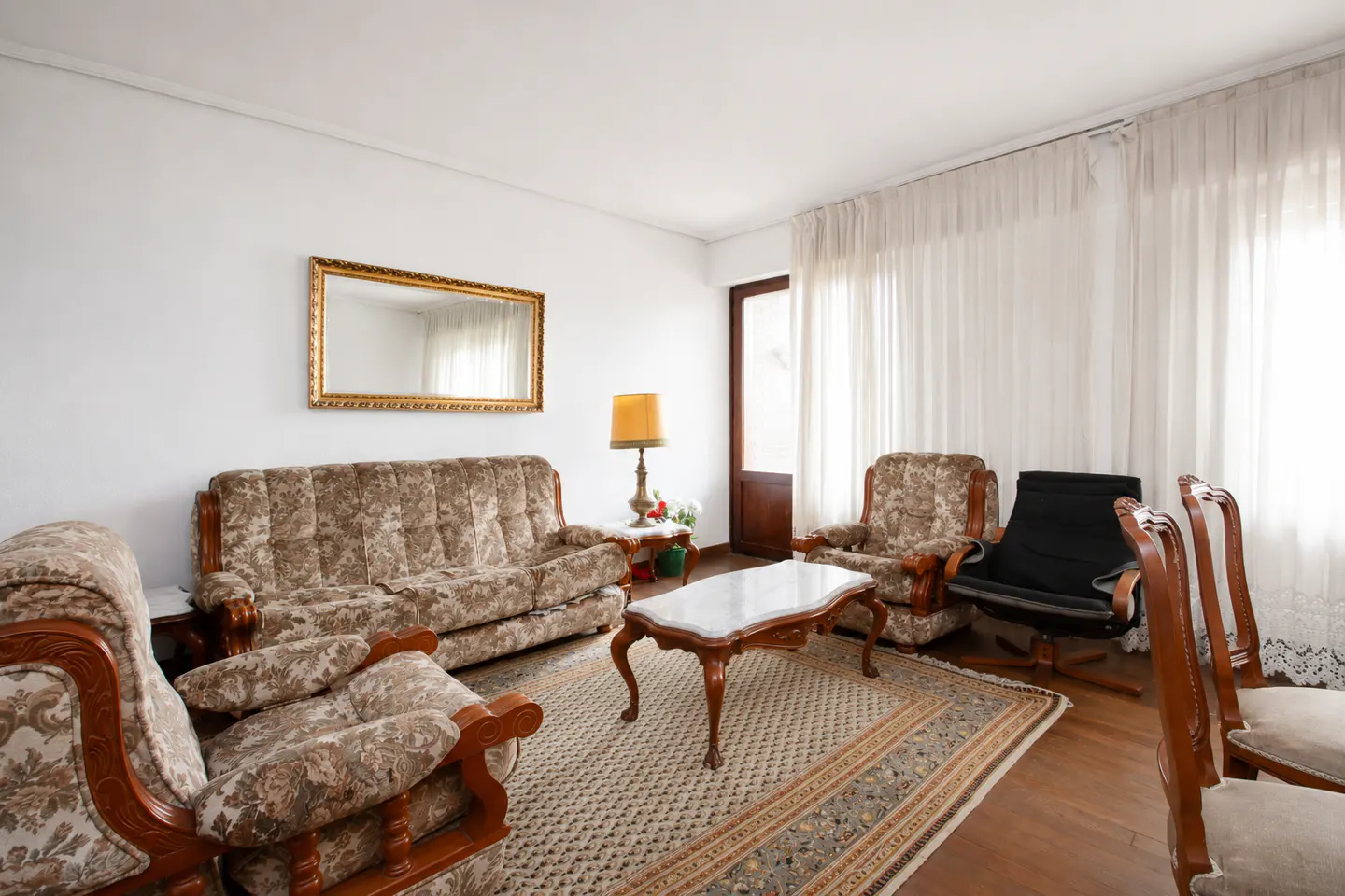 Living room with floral sofa and armchairs, a marble-topped coffee table, and a gold-framed mirror on a white wall.