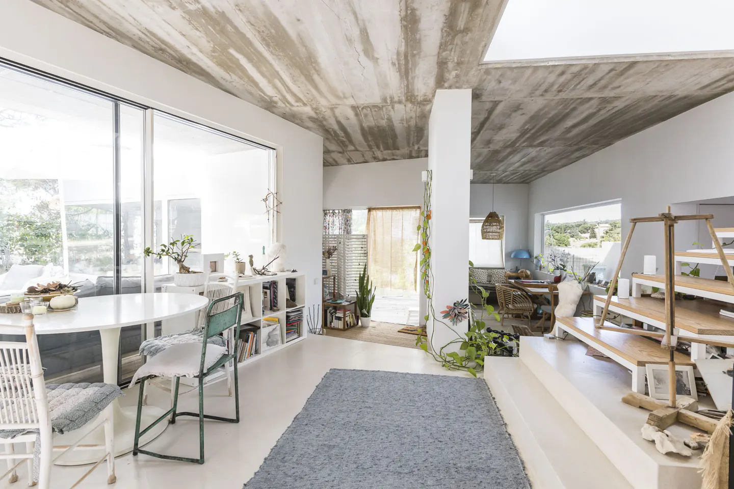 Modern living room with raw concrete ceiling, white walls, dining area, blue rug, and tiered wooden display platform.