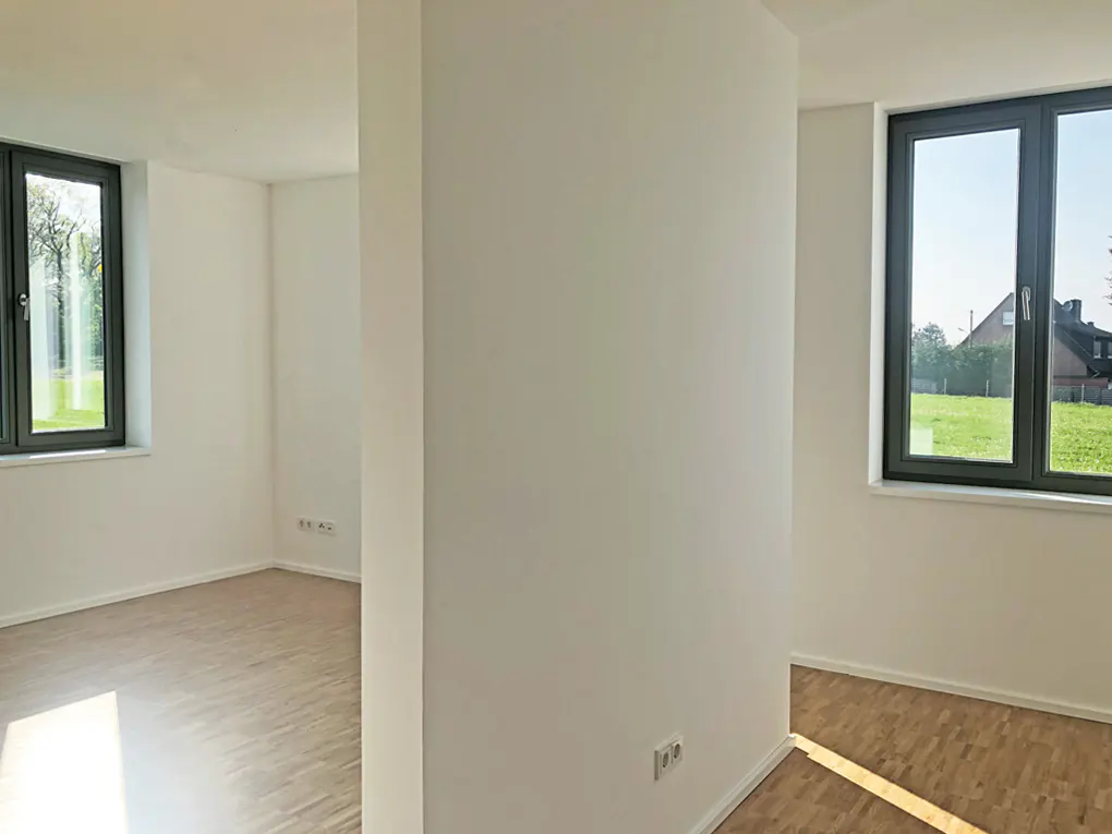 Bright, empty room with light wood floors, white walls, and dark-framed windows overlooking green grass and trees.