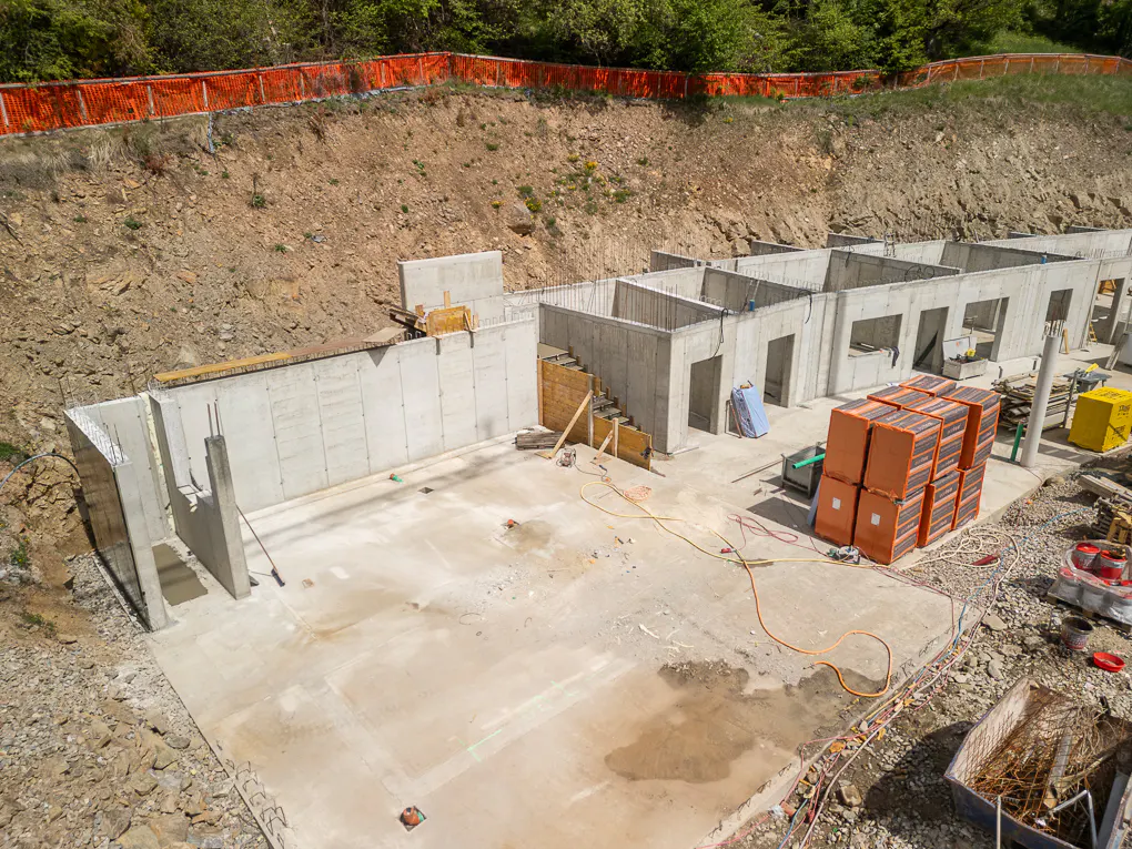 Construction site with concrete foundation and walls. Orange safety fence on a dirt hill in the background.
