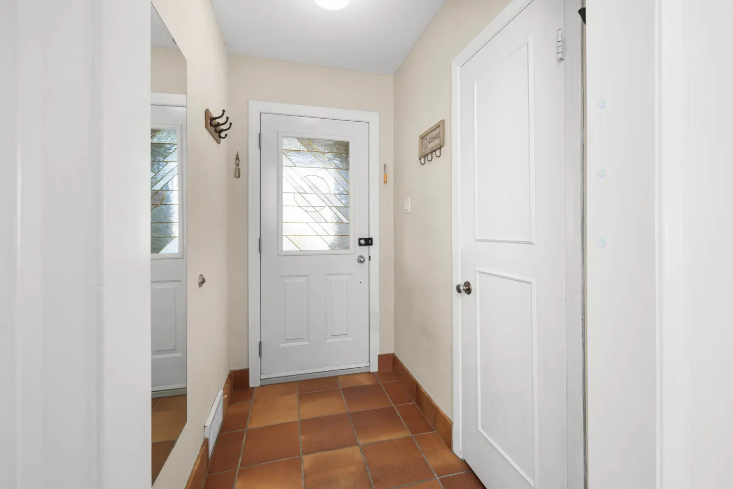 Entryway with terracotta tile floor, white walls, and two white doors. One door has a glass window. Coat hooks are on the wall.
