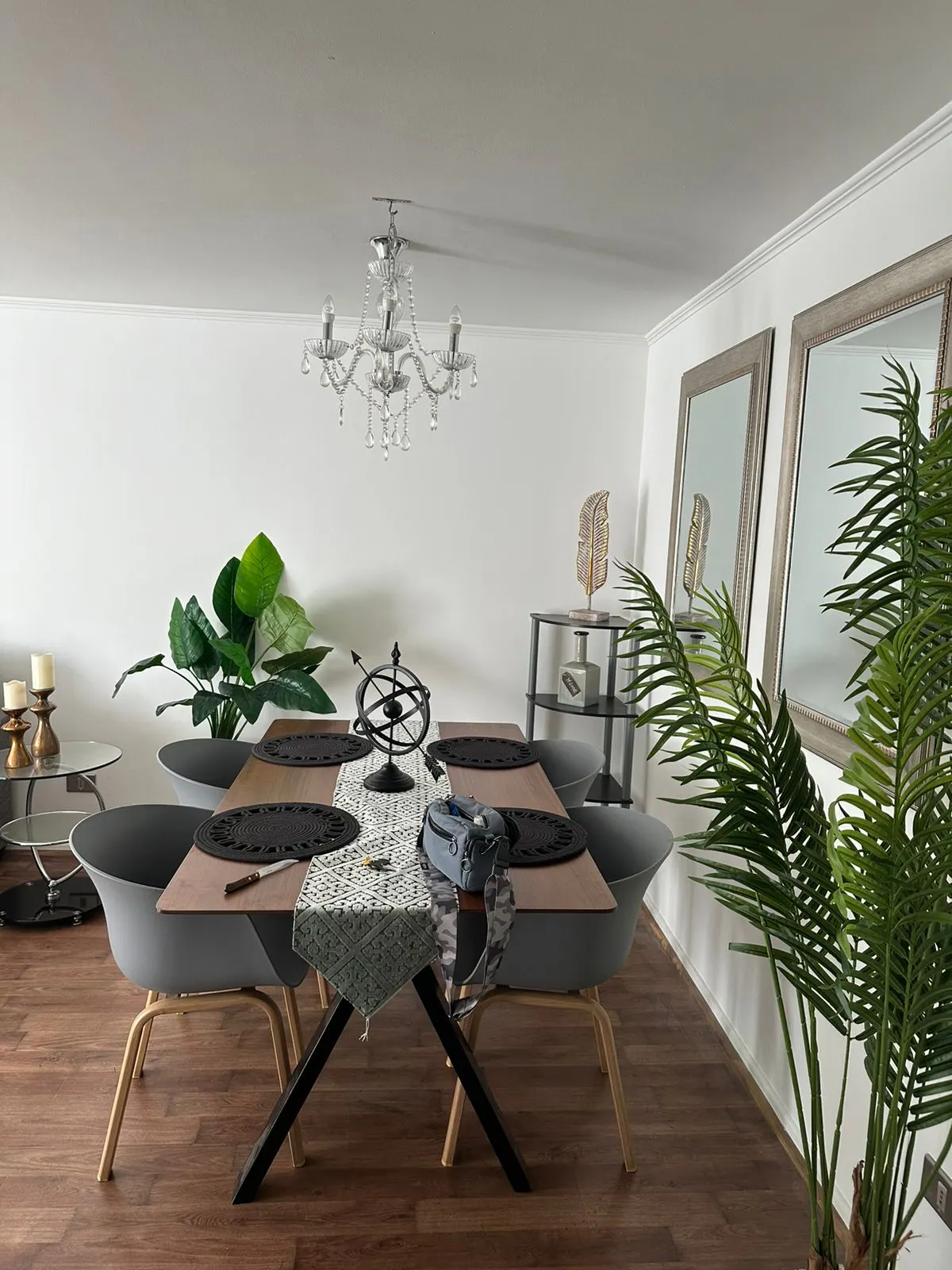 A dining room with a wooden table, gray chairs, and a crystal chandelier. Two mirrors hang on the white wall.