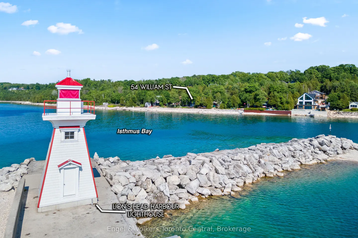 Aerial view of Lion's Head Harbour Lighthouse, a white structure with a red roof, on a rocky pier extending into turquoise water. Trees and a house are visible in the background.