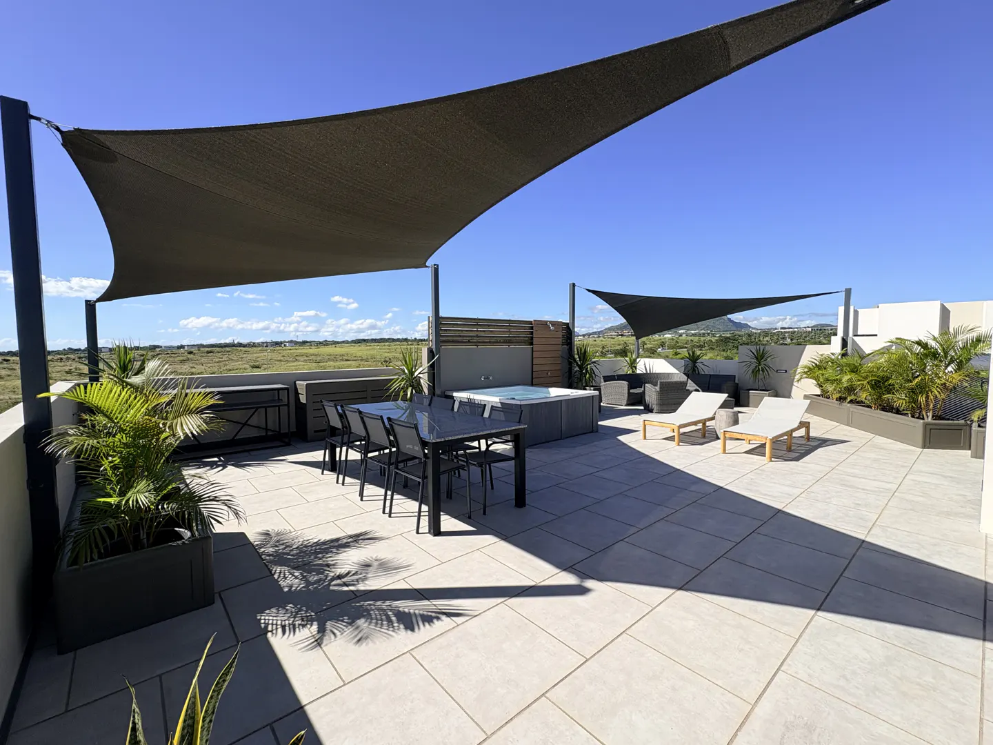 Rooftop patio with dining table, hot tub, lounge chairs, and shade sails under a blue sky.
