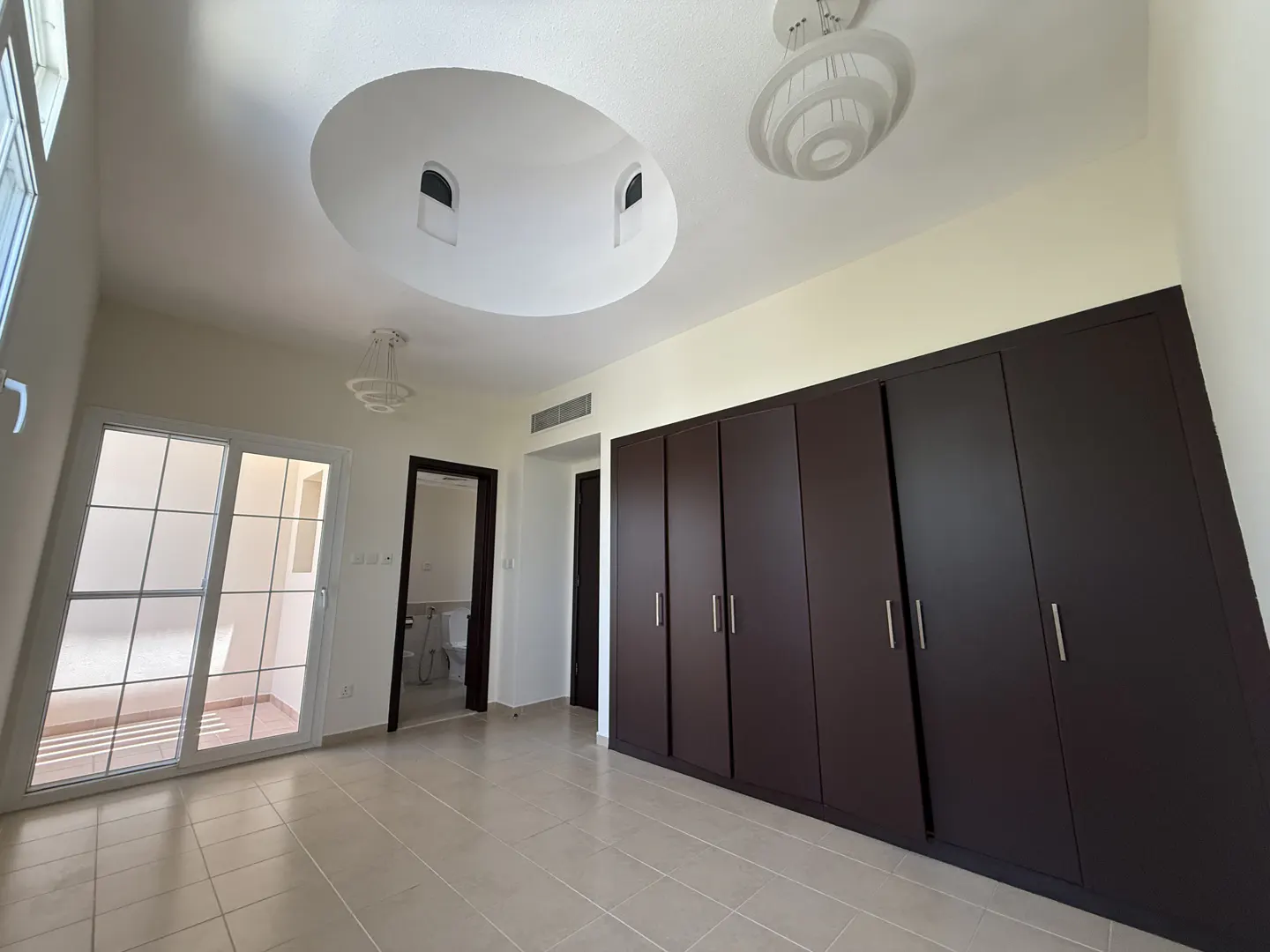 A bright, empty bedroom with a large dark brown wardrobe, sliding glass doors, and a unique ceiling design.