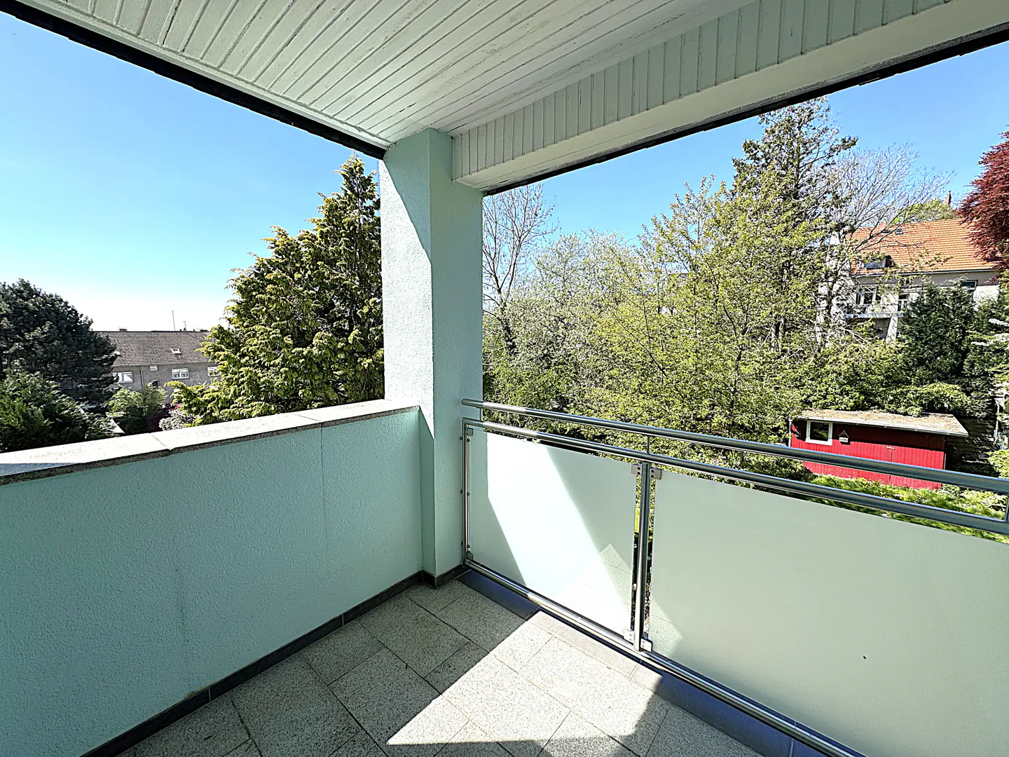 A light blue balcony with a metal railing overlooks lush green trees and a clear blue sky.