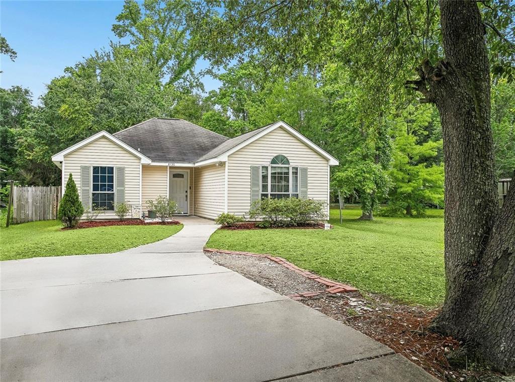 Beige single-story house with gray shutters, a concrete driveway, and green lawn surrounded by trees.