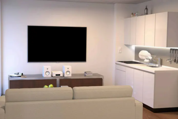 Living room with a large black TV, white speakers, and a beige sofa. The kitchen has white cabinets and stainless steel appliances.