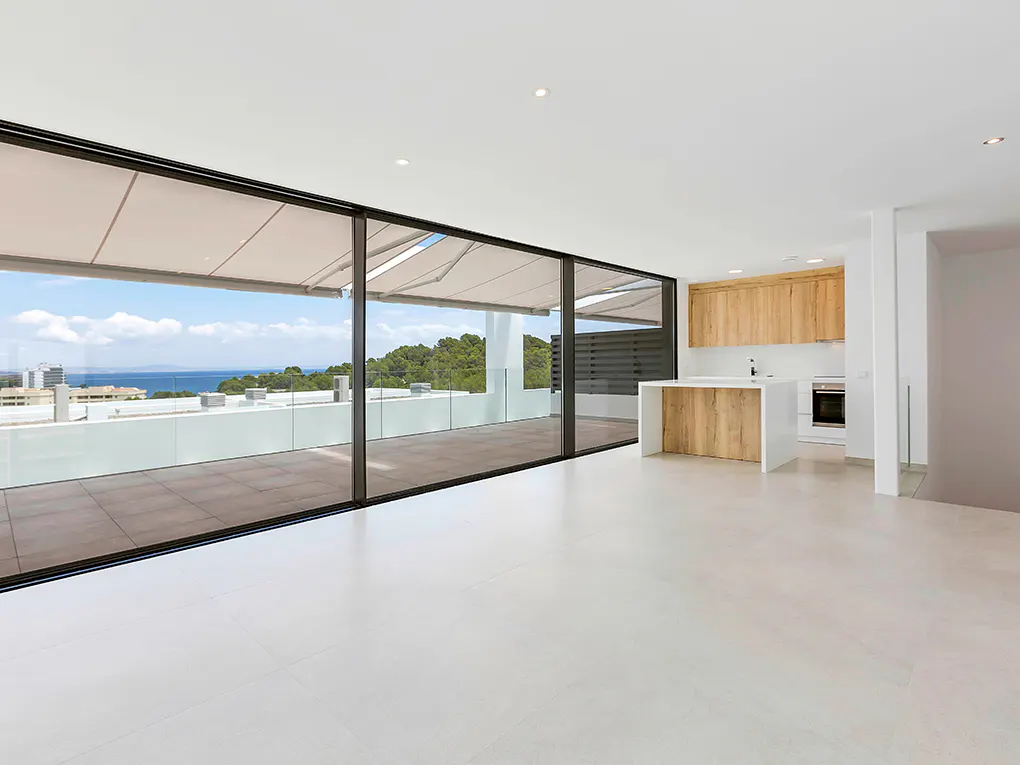 Bright, modern apartment with a large sliding glass door to a balcony overlooking the ocean and trees. The kitchen has wood cabinets and a white island.