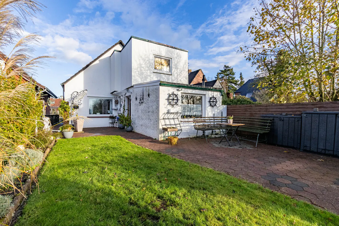Exterior view of a white house with a green lawn, brick patio, and outdoor seating. Blue sky with clouds in the background.